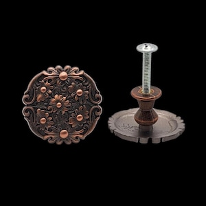 May include: Two decorative cabinet knobs with a floral design. One knob is fully assembled with a screw, the other is a standalone knob. The knobs are bronze and copper-colored, with a dark background.