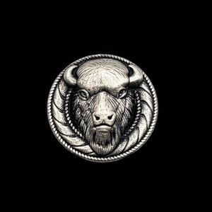 May include: A silver-colored, circular button with a detailed relief of a bison's head. The bison is centered, with horns and a textured face. The button's edge features a rope-like design, set against a black background.