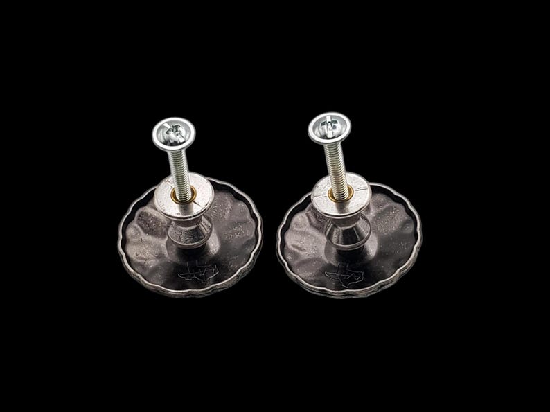 Może przedstawiać: Dwa srebrne gałki do szafek z teksturowaną, ciemnoszarą powierzchnią i falistą krawędzią. Każda gałka ma srebrną śrubę i mały zarys Teksasu. Gałki są ustawione na czarnym tle.