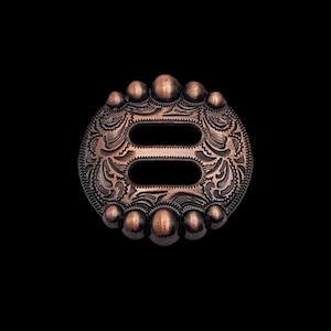 以下が含まれることがあります： 装飾的な彫刻が施された、銅色の金属製のコンチョ。ダブルスロットの開口部があります。コンチョは丸みを帯びた縁に小さな隆起した突起があります。