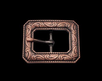May include: A copper-toned belt buckle with an ornate, engraved design. The buckle has a rectangular shape with a smooth, black metal center.