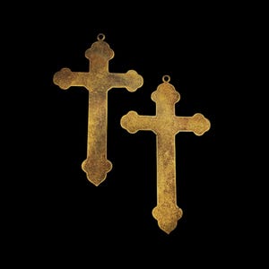 May include: Two gold-toned metal crosses with a distressed finish. The crosses are ornate and have a slightly rounded shape.