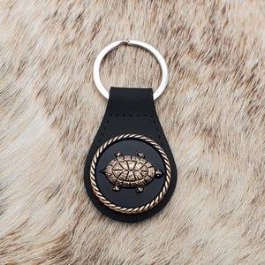 May include: Black leather keychain with a silver key ring. The keychain features a circular black emblem with a gold-colored turtle design, framed by a gold rope-like border. The background is a textured, light brown surface.