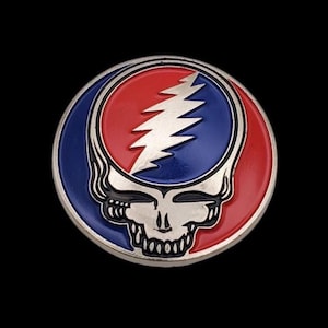 May include: A round, metal pin featuring a skull with a lightning bolt and a red and blue color scheme. The pin has a silver border and is set against a black background. The skull has a smiling expression.