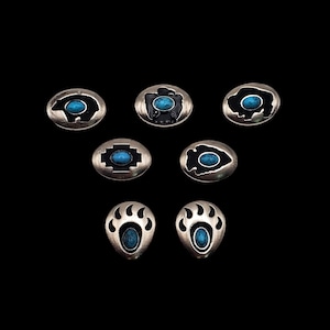 Può includere: Sette bottoni in metallo color argento con pietre turchesi. Ogni bottone presenta un diverso design nativo americano, tra cui un orso, un'aquila, un bisonte, un'impronta di zampa, un simbolo e una punta di freccia.