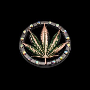May include: A black metal circle with a green and copper marijuana leaf design. The circle is decorated with iridescent rhinestones.