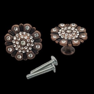 May include: Two antique copper-toned cabinet knobs with a floral design. Each knob features a circular base with a raised centre surrounded by a ring of rhinestones. The knobs are attached to screws.