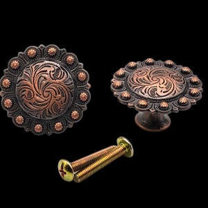 Op de afbeelding: Twee koperkleurige kastknoppen met een decoratief bloemmotief. De knoppen hebben een ronde basis met een verhoogd midden en zijn omgeven door een ring van kleine, decoratieve accenten. De knoppen worden geleverd met twee schroeven.