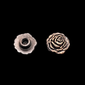 Pode incluir: Dois botões de metal em forma de rosa, um com acabamento em cobre e outro com acabamento em prata. O botão de cobre tem um design de rosa em relevo, enquanto o botão de prata tem uma superfície plana com um pequeno furo no centro.