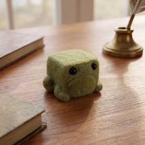 Può includere: Una piccola statuetta a forma di cubo realizzata in feltro verde oliva. L'oggetto presenta due occhi circolari neri lucidi e una semplice linea curva che rappresenta una bocca, conferendogli un aspetto distintivo, squadrato e animale.