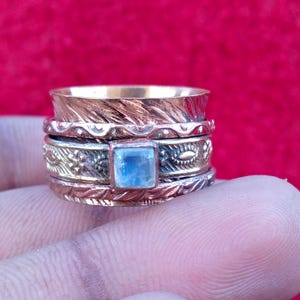 Peut inclure: Une bague rotative multi-anneaux en cuivre et laiton, orn&eacute;e d'un cabochon central en pierre de lune de forme carr&eacute;e. La bague pr&eacute;sente des motifs gaufr&eacute;s complexes, incluant des textures de feuilles et des lignes ondul&eacute;es autour de la pierre.