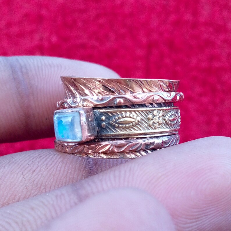 Peut inclure: Une bague rotative multi-anneaux en cuivre et laiton, orn&eacute;e d'une pierre de lune rectangulaire centrale aux reflets bleut&eacute;s. La bague pr&eacute;sente des motifs gaufr&eacute;s complexes, incluant des d&eacute;tails floraux et des bords textur&eacute;s sur les anneaux.