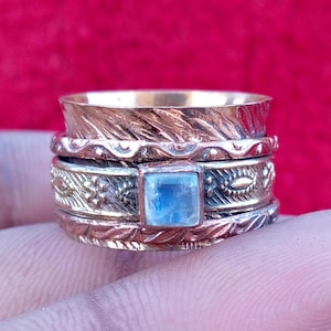 Peut inclure: Une large bague rotative en métaux mixtes, composée d'une bande cuivrée à motif de feuilles texturées et d'une bande centrale argentée ornée d'une pierre de lune bleue carrée et irisée, sertie et décorée de motifs floraux.