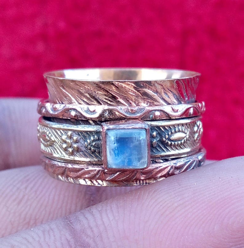 Peut inclure: Une large bague rotative en m&eacute;taux mixtes, compos&eacute;e d'une bande cuivr&eacute;e &agrave; motif de feuilles textur&eacute;es et d'une bande centrale argent&eacute;e orn&eacute;e d'une pierre de lune bleue carr&eacute;e et iris&eacute;e, sertie et d&eacute;cor&eacute;e de motifs floraux.