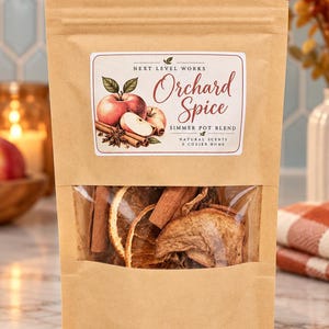 Könnte beinhalten: Ein brauner Papierbeutel mit einer Orchard Spice Simmer Pot Mischung. Das Etikett zeigt eine Illustration eines Apfels, Zimtstangen und Sternanis mit dem Text Next Level Works Orchard Spice Simmer Pot Blend Natural Scents A Cozier Home.