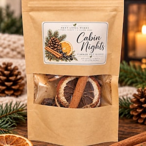 Op de afbeelding: Een bruine papieren zak met een simmer pot blend. Het etiket bevat de tekst "Next Level Works", "Cabin Nights" en "Simmer Pot Blend" met illustraties van een dennenappel, sinaasappelschijfje en kaneelstokjes. Het doorzichtige venster toont gedroogde sinaasappelschijfjes en kaneelstokjes.