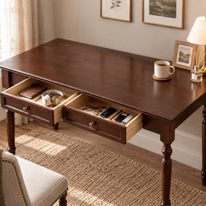 May include: A dark brown wooden desk with four turned legs and two open drawers. The left drawer contains a notebook and headphones, while the right drawer holds eyeglasses and small electronic items. The desk surface features a white mug and a lamp.