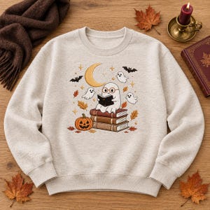 Puede incluir: Una sudadera de cuello redondo en color beige jaspeado con un gráfico central de un fantasma con gafas, leyendo un libro mientras está sentado sobre una pila de tres libros. El diseño incluye una calabaza, tres fantasmas pequeños, una luna creciente, murciélagos y hojas de otoño.