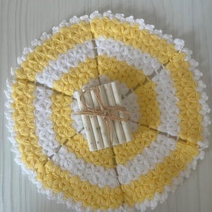 Puede incluir: Un tapete redondo de ganchillo con anillos concéntricos de hilo amarillo y blanco en un patrón de punto texturizado en forma de estrella. Centrado en el tapete hay una pila rectangular de cuatro pastillas de jabón blanquecinas atadas con cordel.