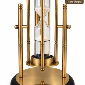 Könnte beinhalten: Eine Sanduhr aus rohem Messing mit einer zentralen Glaskammer, die mit weißem Sand gefüllt ist, getragen von einem vertikalen Metallrahmen mit zylindrischen Säulen und einem runden Sockel. Die Struktur umfasst einen schwarzen Ring am Boden und einen kleinen, mit Raw Brass beschrifteten Kreis.