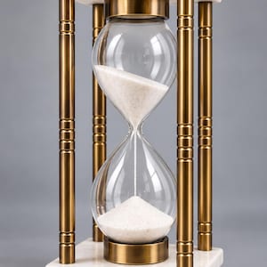 May include: An hourglass featuring a clear glass bulb filled with white sand, held within a frame of four metallic bronze-colored pillars. The structure is supported by a white marble base and top, each accented with small spherical bronze feet and finials.