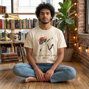 Peut inclure: Un t-shirt à manches courtes de couleur crème avec un motif représentant un chat tigré portant une casquette rouge et des lunettes de soleil, effectuant un dab. Sous le chat, le texte indique : EAT. SLEEP. CODE. PURR. REPEAT. T-shirt en coton.