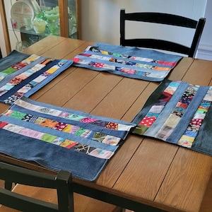 May include: Four denim placemats with colorful patchwork accents are arranged on a wooden table. The placemats are rectangular with a denim border and a central strip of various fabric scraps. A black chair is in the background.