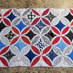May include: A handmade quilt featuring a repeating pattern of overlapping circles and diamond shapes. The quilt showcases a variety of fabrics in red, white, blue, black, and gray. The design includes floral, polka dot, and striped patterns, creating a visually dynamic textile.