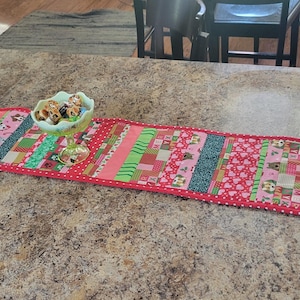 May include: A long, rectangular quilted table runner with a red border and a patchwork design. The patchwork includes various fabrics in shades of pink, green, and blue, with patterns of hearts, stripes, and small squares. A small, decorative bowl sits on top.