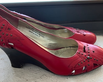 May include: A pair of red leather wedge heels with cut-out designs and studded accents. The shoes have a black wedge heel and a size 7 label inside. The shoes are on a white surface.