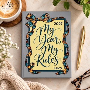 Może przedstawiać: Szary planer w twardej oprawie z żółtym prostokątnym środkiem zawierającym napis "2027 My Year, My Rules" oraz imię "Lucy" ciemnoniebieską kursywą. Ozdobna wstążka z wzorem niebiesko-pomarańczowych skrzydeł motyla otacza tekst.