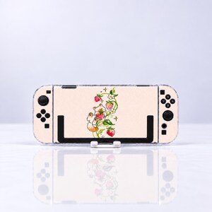 May include: A beige gaming console skin featuring a central illustration of a small cat climbing a green vine with red strawberries and white flowers. The skin covers the back of the handheld device, which has black buttons and joysticks on both sides.