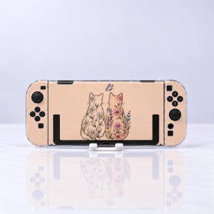 May include: A beige gaming console skin featuring two cats sitting side by side, decorated with floral patterns of white daisies and pink flowers, with a small butterfly above them. The skin is applied to a handheld gaming device with black controllers.