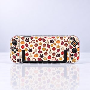 May include: A handheld gaming console featuring a cream-colored skin with a repeating autumn-themed pattern. The design includes small black cat faces, orange pumpkins, red apples, brown acorns, yellow star-shaped cookies, candles, and maroon leaves.