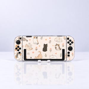 May include: A handheld gaming console featuring a cream-coloured protective case decorated with a repeating pattern of various illustrated cats, small flowers, and paw prints. The device includes black buttons, joysticks, and a black central stand.