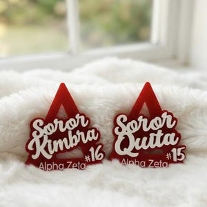 Peut inclure: Deux ornements triangulaires en acrylique rouge avec du texte en écriture blanche. L'ornement de gauche indique Soror Kimbra #16 Alpha Zeta, et celui de droite indique Soror Quita #15 Alpha Zeta. Les deux pièces ont une finition lisse et brillante.