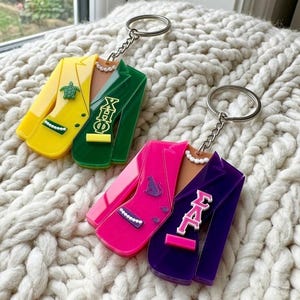 May include: Two acrylic keychains shaped like blazers. The first is yellow and green with a turtle emblem and Greek letters XHΦ. The second is pink and purple with a panther emblem and Greek letters ΣΛΓ. Both feature white pearl-like accents.