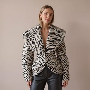Puede incluir: Una chaqueta estructurada con un estampado de cebra en negro y crema. La prenda presenta un cuello chal ancho y sobredimensionado, mangas largas y una cintura entallada que se ensancha ligeramente en las caderas. La tela tiene textura acolchada.
