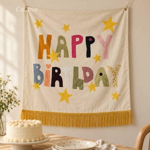 May include: A cream-colored fabric wall hanging featuring the text HAPPY BIRTHDAY in multicolored, patterned fabric letters. The banner is decorated with yellow fabric stars and finished with a long yellow fringe along the bottom edge.