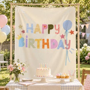 May include: A cream-colored fabric backdrop featuring the text HAPPY BIRTHDAY in multi-colored, patterned fabric letters. The banner is decorated with fabric stars, a crown, and a blue balloon. It is displayed on a wooden frame stand.