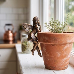 May include: A bronze-colored fairy figurine with detailed wings and hair, posed as if leaning against the rim of a terracotta flower pot. The fairy has a smooth metallic finish and is attached to the side of the round, weathered clay pot.