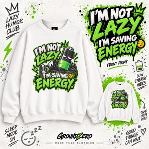 Può includere: Una felpa bianca a girocollo con la grafica di una batteria che riposa e il testo I'M NOT LAZY I'M SAVING ENERGY. Il design utilizza colori verde brillante e nero con scritte in stile graffiti e un'emoji con faccia assonnata.