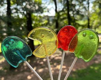 May include: Four colorful lollipops shaped like ghosts and Pac-Man. The lollipops are on white sticks and are arranged in a semi-circle.