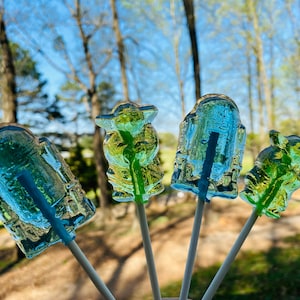 May include: Four Star Wars character lollipops on sticks. The lollipops are clear blue and green and shaped like Yoda, R2-D2, a Stormtrooper, and a character from the Mandalorian.
