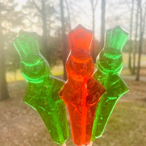 May include: Three candy lollipops on sticks. One is red and the other two are green. The lollipops are shaped like people.