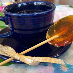 May include: A blue teacup and saucer with a gold ribbon tied around a wooden stick holding a clear amber lollipop.