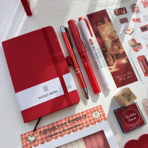 May include: A red A6 pocket book with an elastic band, a red metallic ballpoint pen, a red fountain pen, a white highlighter, a bookmark with a teddy bear illustration, a wooden stamp, and a red ink pad with the text Bentota House.