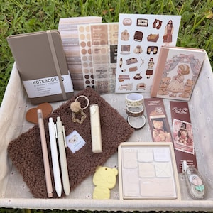 Peut inclure: Un ensemble de papeterie comprenant un carnet A6 rigide taupe, une pochette en peluche marron, trois stylos, un objet jaune en forme de panda, des planches d'autocollants, du ruban adhésif washi, un petit livre illustré, des marque-pages et un bloc-notes.