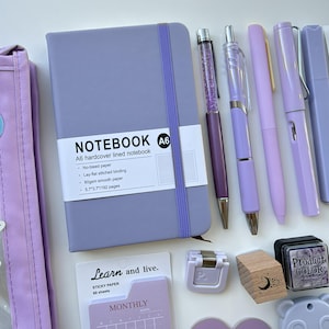 Könnte beinhalten: Ein lavendelfarbenes A6-Hardcover-Notizbuch mit Gummiband, vier lila Stiften, einem Block monatlicher Haftnotizen mit der Aufschrift Learn and live, einem kleinen lila Hefter, einem Holzblock mit Mondsymbol und einem Product Color Stempelkissen.