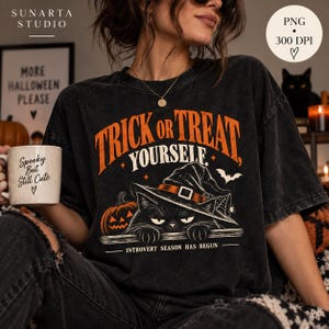 May include: A black acid-wash t-shirt featuring an orange and white graphic of a black cat wearing a witch hat, a pumpkin, and a bat. The text reads TRICK OR TREAT YOURSELF and INTROVERT SEASON HAS BEGUN. A white mug with the text Spooky But Still Cute.